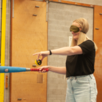 A lady blindfolded holding a baseball bat and baseball, positioned ready to hit.