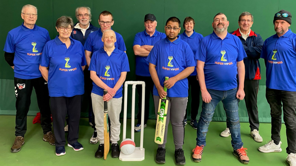 A group of people in an indoor sport arena wearing blue tshirts, one is holding a cricket bat and they are stood behind a wicket.