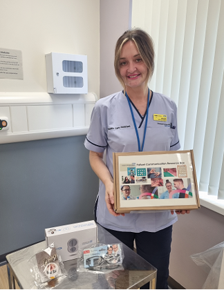 A hospital clinical team member in a lilac tunic holding a photoframe and standing next to a box of accessories to assist with accessibility.