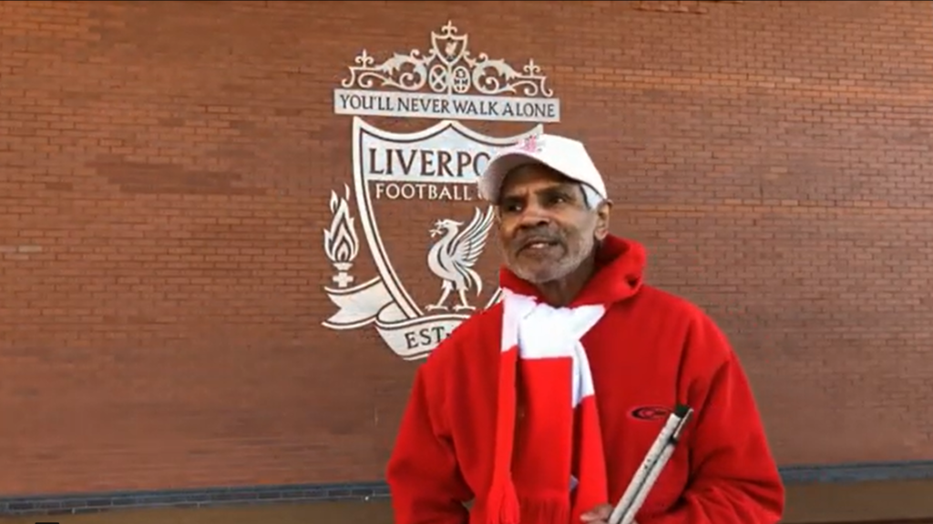 Image of a gentleman wearing a red jacket, a red and white scarf stood in front of the Liverpool FC crest at Anfield and holding a white cane
