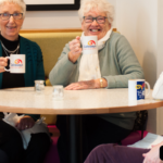 A group of people smiling, holding branded cups with the Galloways logo on. The bottom right corner has a caption in white writing on top of a black box saying," brews up". The top left corner has Galloways logo with an orange semi-circle behind.