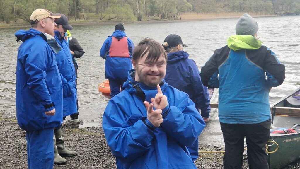 An image of Dan posing with his hands and a big smile on his face stood on a stony bay in the Lake District next to a group of people looking out on the water.