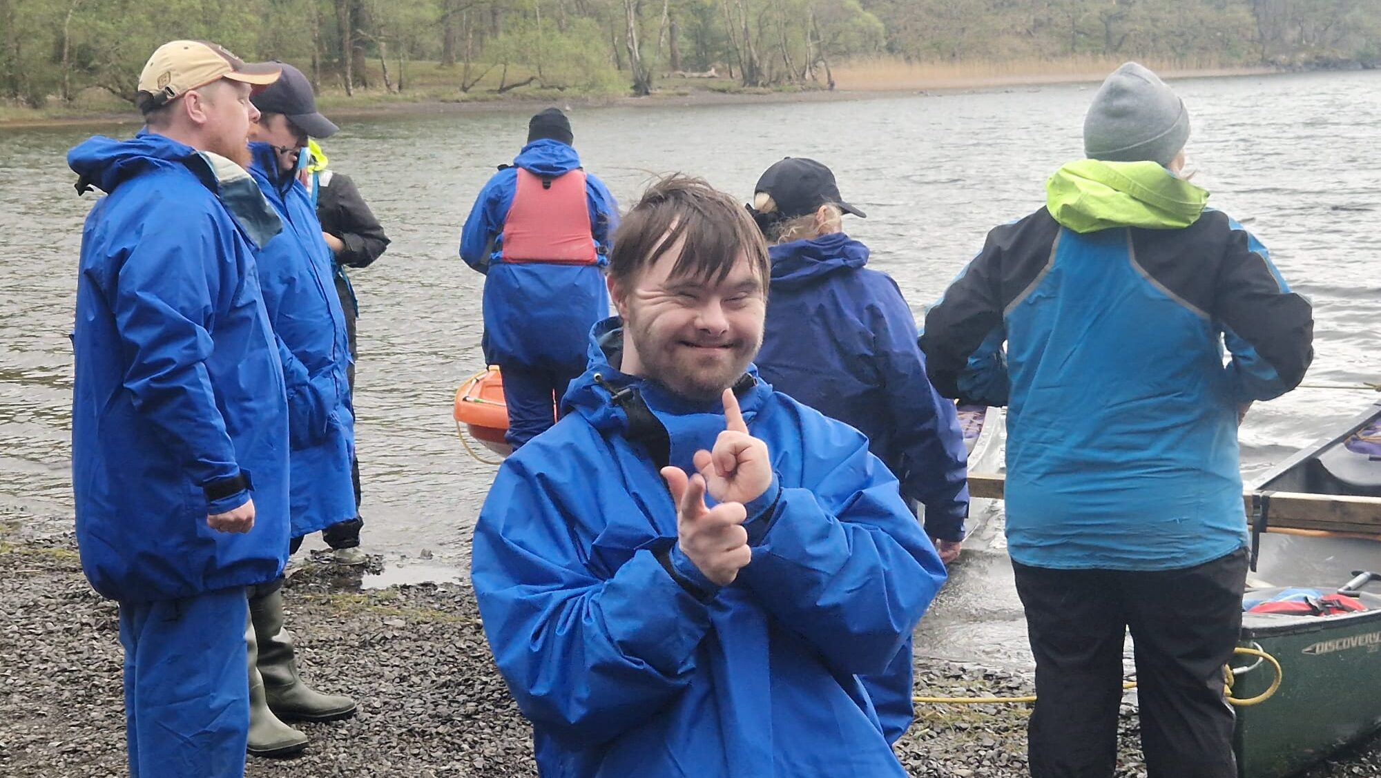 An image of Dan posing with his hands and a big smile on his face stood on a stony bay in the Lake District next to a group of people looking out on the water.