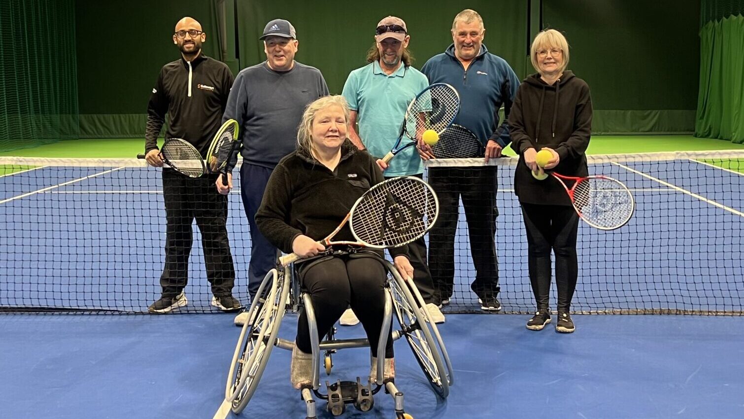 Image of a group of people holding tennis rackets on a tennis court. One person at the front is in a wheelchair.