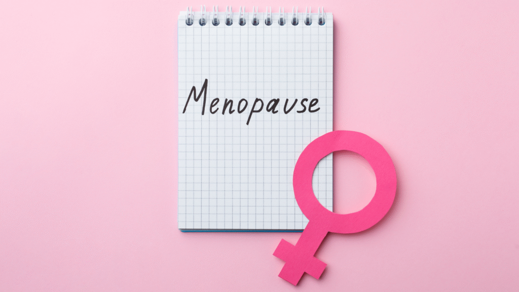 A light pink background with a white notepad in the centre with the word "menopause" written in black handwriting, with the universal symbol for female situated diagonally in the corner.