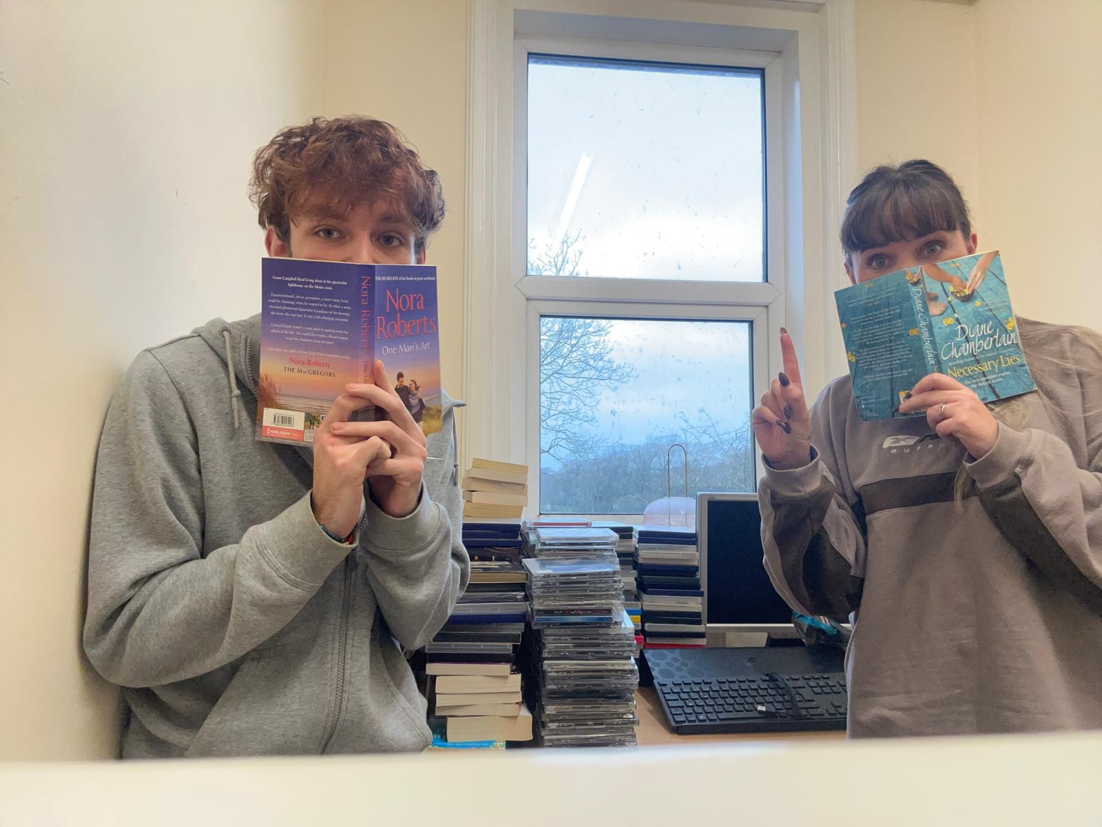 2 people hiding their faces behind books with a stash of books, CDs and DVDs behind them.