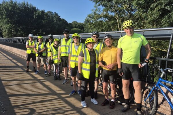 Alt text: A group of cyclists wearing helmets and high-visibility clothing stand together on a bridge surrounded by trees. A blue tandem bike is visible beside the group on a bright, sunny day.