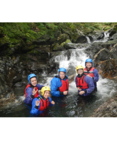Five people wearing helmets, life jackets and waterproof clothing smile and wave while standing in a rocky stream beneath a waterfall.