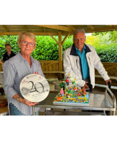A woman and man stand beside a colourful cake celebrating 20 years of Galloways Water Park. The woman holds a commemorative plaque, with a wooden gazebo and greenery behind them.