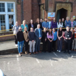 Image of the Galloways team stood outside of the headquarters at Howick House, an old red stone brick building. There is a white cr parked to the left and around 20 people gatehred in the centre looking towards the camera smiling.