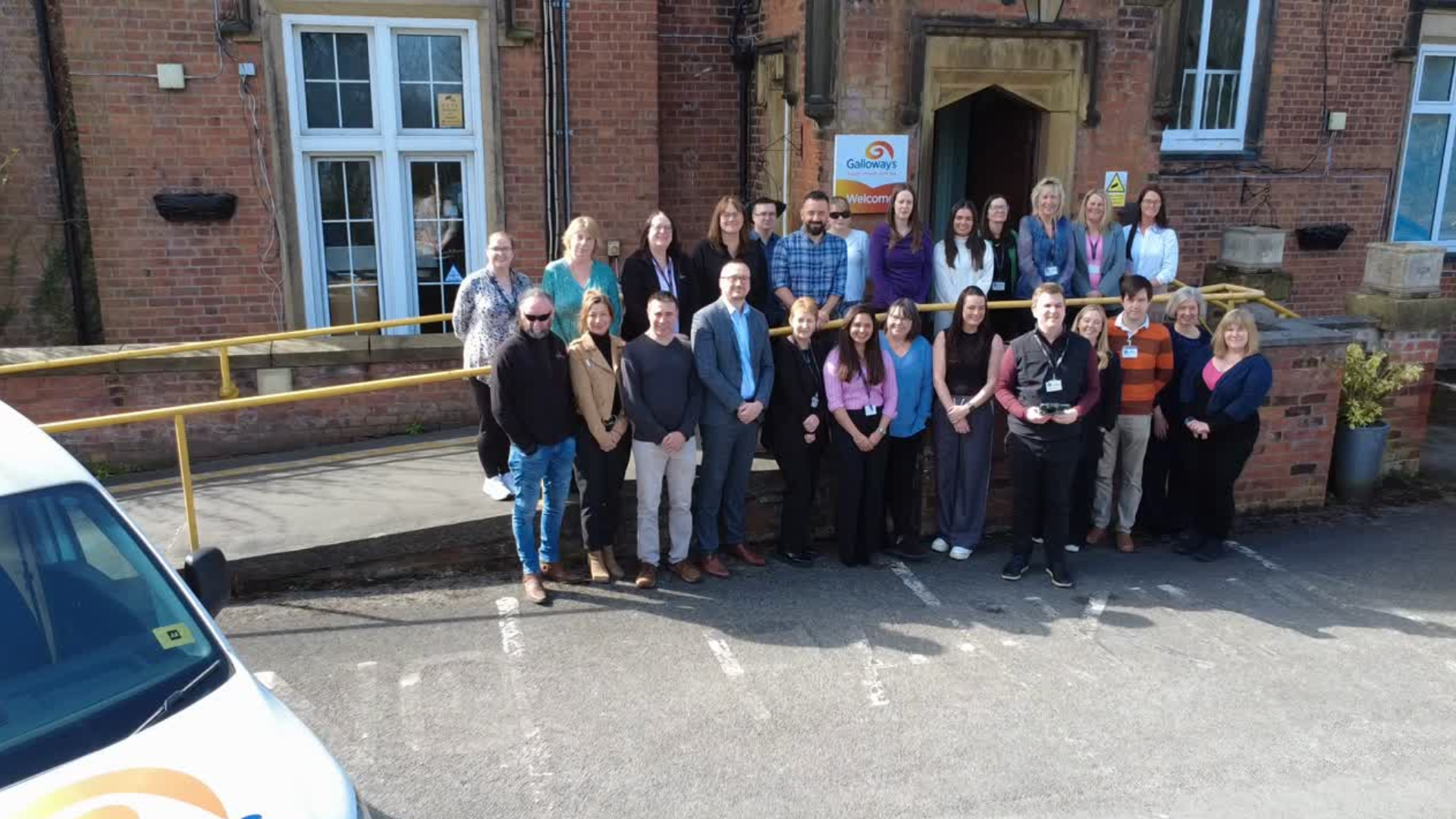 Image of the Galloways team stood outside of the headquarters at Howick House, an old red stone brick building. There is a white cr parked to the left and around 20 people gatehred in the centre looking towards the camera smiling.